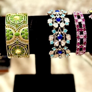 May include: An assortment of decorative bracelets in various styles and colors. The bracelets include designs with green, yellow, blue, and pink gemstones and floral accents. The bracelets are displayed on a black stand.