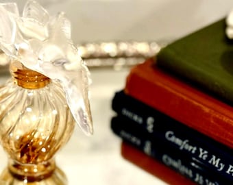 May include: A clear glass perfume bottle with a twisted, ribbed body and a gold-toned neck. The stopper is a translucent, sculpted floral or leaf-like design. Beside the bottle are three stacked books with green, red, and black covers.