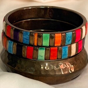 May include: A stack of three bangles. The bottom bangle is dark, hammered metal. The two top bangles feature alternating vertical rectangular inlays in vibrant colors including red, orange, green, blue, purple, and white, separated by dark metal spacers.