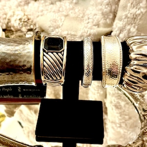 May include: A collection of silver and gold-toned bracelets displayed on a black stand. The bracelets feature various designs, including hammered textures, geometric patterns, and a black stone accent. The bracelets are displayed on a black stand.