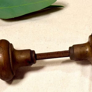 May include: Antique bronze door knob with a threaded rod connecting two rounded knobs. The knobs have a warm, aged patina. A green leaf and stem are in the background.