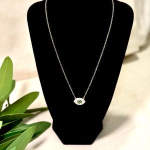 May include: A silver-toned necklace featuring an eye-shaped pendant. The pendant has a green center and is surrounded by small, clear stones. The necklace is displayed on a black mannequin.