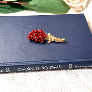 May include: A vintage brooch featuring a bouquet of red flowers with gold-tone stems and leaves, resting on a navy blue book. The book has the title "Comfort Ye My People" and the publisher "Macmillan".