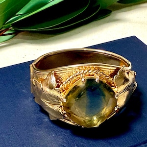 May include: A gold-toned hinged bangle bracelet with a large, faceted, yellow-green gemstone. The bracelet features leaf and rope detailing. The gemstone is approximately 1 inch in diameter. The bracelet is set on a blue surface.