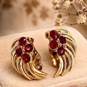 May include: A pair of vintage gold-toned clip-on earrings. Each earring features a cluster of five round, deep red gemstones set within a swirling, textured gold design. The earrings are displayed on a soft, beige surface.