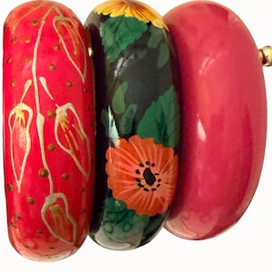 May include: A set of three wooden bangles. One is red with gold floral designs, the second is black with a red flower and green leaves, and the third is a solid red. The bangles are connected by a gold-colored metal piece.