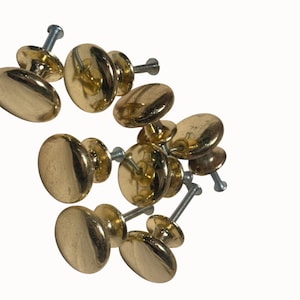 May include: A collection of gold-colored cabinet knobs with a rounded, oval design. Each knob has a threaded screw for attachment. The knobs are arranged against a white background, suggesting a hardware or home decor theme.