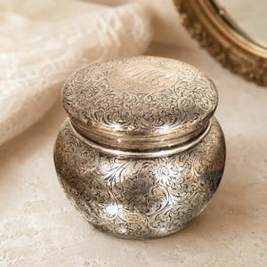 May include: A silver-toned, antique jar with a lid. The jar and lid are covered in an intricate floral pattern. The lid has an engraved monogram. The jar is round with a slightly bulbous body. The background includes a mirror and lace.