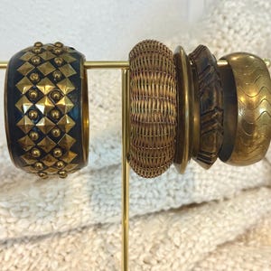 May include: A collection of bangles displayed on a gold stand. The bangles include a dark blue and gold studded bangle, a woven rattan bangle, and several gold and brown bangles with carved and wave designs.