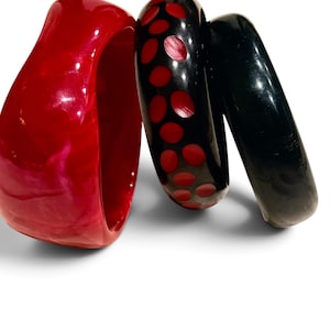 May include: A set of three bangle bracelets. One is a wide, glossy red bracelet with a marbled texture. The second is a black bracelet featuring a pattern of recessed red circular dots. The third is a smooth, solid black bangle bracelet.