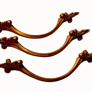 May include: A set of four antique-style drawer pulls. Each pull is a curved, bronze-colored metal with ornate leaf and ball detailing at each end. The pulls are arranged against a white background.