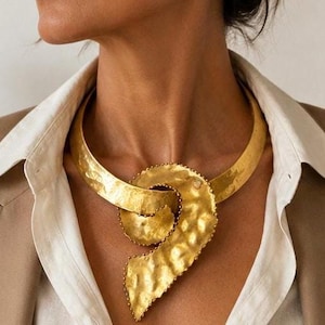 May include: A gold-toned metal collar necklace featuring a wide, hammered, textured band that curves into a prominent, swirling, organic-shaped pendant at the center. The surface of the metal has a matte, dimpled finish with defined edges.