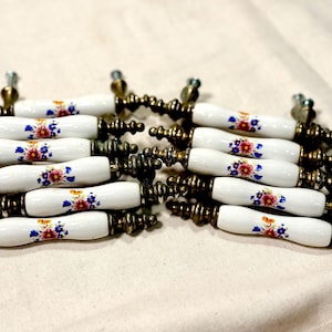 May include: A collection of white ceramic cabinet pulls with a floral design in red, blue, and yellow. The pulls have brass-colored metal hardware and are arranged in two rows on a neutral surface.