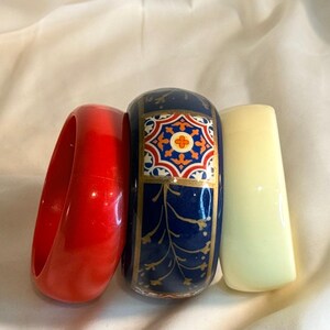 May include: Three bangle bracelets arranged side by side. The left bracelet is a solid, glossy red. The center bracelet is dark blue with a gold-outlined geometric tile pattern and gold leaf-like accents. The right bracelet is a solid, glossy cream.