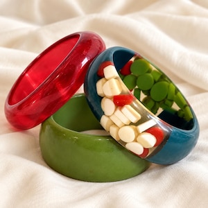 May include: Three bangle bracelets stacked together. One is a translucent red, one is a solid matte olive green, and the third is a dark teal resin bangle embedded with small, scattered white, red, and green geometric shapes and circular pieces.