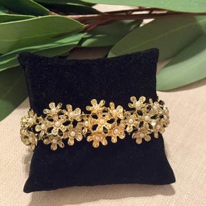 May include: A gold-toned bracelet with a floral design, featuring small pearl accents. The bracelet is displayed on a black velvet cushion, with green leaves in the background. The bracelet's design is intricate and detailed.