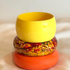 May include: A stack of three bangle bracelets. The top bangle is solid bright yellow. The middle bangle features a mottled, multi-colored pattern of red, yellow, and black. The bottom bangle is a solid, vibrant orange. Each bracelet is smooth.
