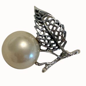 May include: A silver-toned brooch featuring a large, lustrous pearl and a detailed leaf design. The leaf has an open, lattice-like pattern, adding a delicate touch to the jewelry.