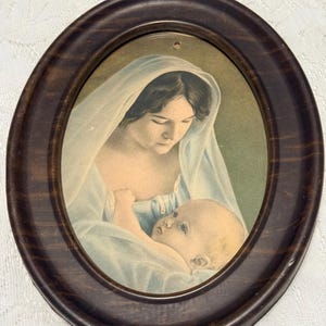 May include: An oval-shaped, framed print depicting a woman in a white veil cradling a baby. The frame is dark brown, and the print features soft colors of blue, white, and skin tones. The artwork is a religious depiction.
