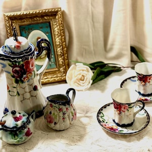 May include: A porcelain tea set featuring a tall teapot, a lidded sugar bowl with a small wooden spoon, a creamer, and two teacups with matching saucers. Each piece is decorated with hand-painted red roses, green leaves, and a gold lattice pattern.