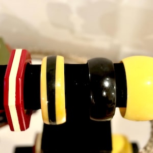 May include: Four colorful bangles displayed on a black stand. The bangles include a red and white striped, a yellow and black striped, a solid black, and a solid yellow bangle. The bangles are made of a glossy material.