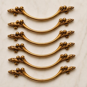May include: Six matching curved drawer pulls in a brushed antique brass finish. Each handle features a central arch with decorative leaf-shaped ends and small spherical accents at the mounting points, arranged in a vertical column.