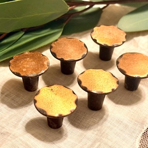 May include: Six decorative cabinet knobs with a mushroom-like design. The knobs have a bronze-colored stem and a scalloped, golden-yellow top. Green leaves are visible in the background.