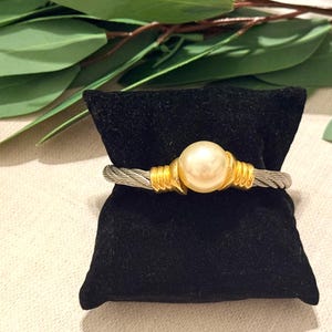 May include: A silver and gold bracelet with a large, round, cream-colored pearl at the center. The bracelet is displayed on a small, black velvet pillow. Green leaves are in the background.