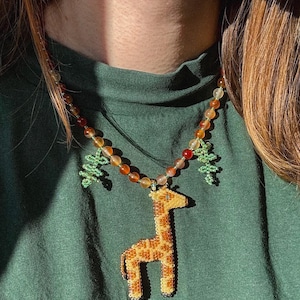 May include: A beaded necklace featuring a giraffe pendant with orange and yellow beads. The giraffe pendant is tan with brown spots. The necklace also has green beaded tree charms and orange and brown beads.