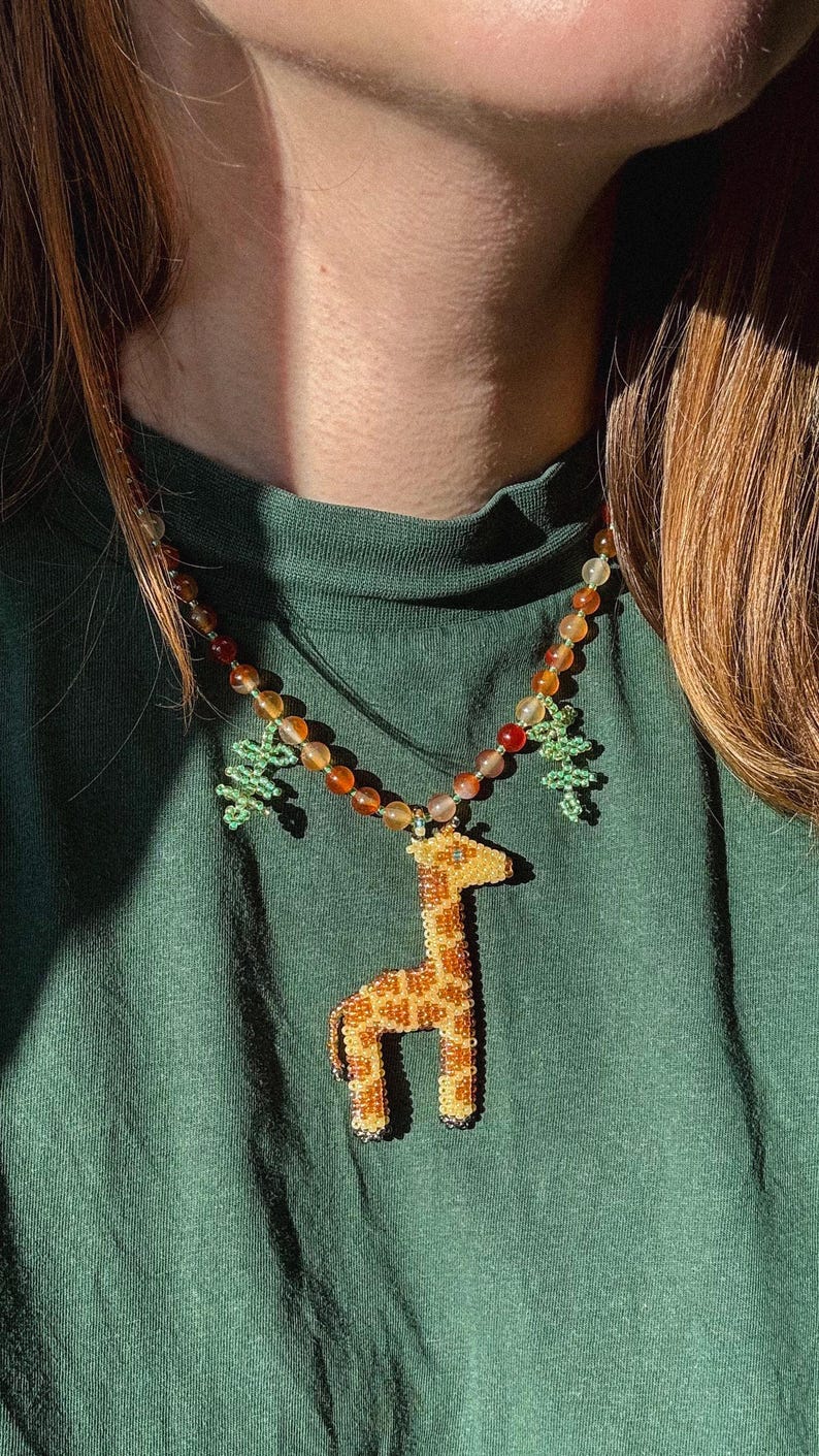 May include: A beaded necklace featuring a giraffe pendant with orange and yellow beads. The giraffe pendant is tan with brown spots. The necklace also has green beaded tree charms and orange and brown beads.