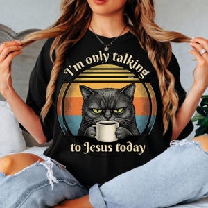 Peut inclure: T-shirt noir avec un chat noir grincheux tenant une tasse à café. Le texte dit "I'm only talking to Jesus today" dans un design circulaire avec un fond de coucher de soleil rétro. Un vêtement décontracté.