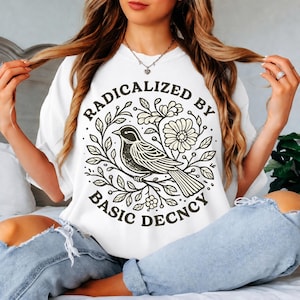 Pode incluir: Camiseta branca com um gráfico circular com um pássaro empoleirado em um galho, cercado por folhas e flores. O texto "RADICALIZED BY BASIC DECNCY" é impresso ao redor do desenho do pássaro. A camisa é de estilo casual.