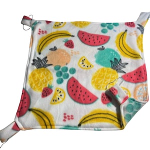 May include: A square, white fleece pet hammock with a colorful fruit pattern. The design includes illustrations of bananas, watermelons, strawberries, pineapples, grapes, and lemons. Metal hooks are attached to the corners for hanging.