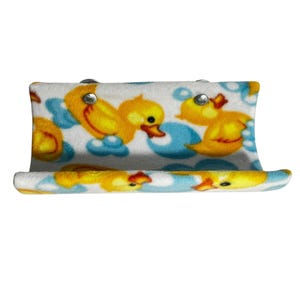 May include: A white, curved, fleece-like shelf with a repeating pattern of yellow rubber ducks on blue clouds. The shelf is secured with silver rivets. The ducks have orange beaks and are in various poses.