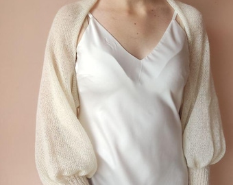May include: Cream-colored knitted bolero with long sleeves and a V-neck. The bolero has a textured knit pattern and is worn over a white dress. The sleeves are gathered at the wrist, creating a slight puff. The background is a solid, light pink.