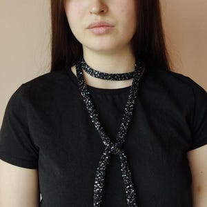 May include: A long, thin, black scarf or neck accessory featuring a dense, shimmering texture created by numerous small, reflective sequins. The accessory is wrapped around the neck and hangs down the chest in a simple, knotted style.
