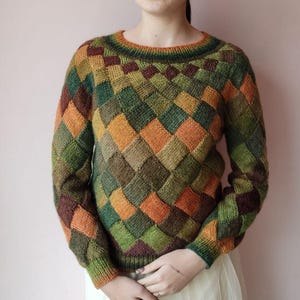 Può includere: Un maglione lavorato a maglia con un motivo geometrico nei toni del verde, arancione e marrone. Il maglione ha un girocollo e maniche lunghe. Il design presenta un motivo a rombi ripetuto. La modella indossa una gonna bianca.