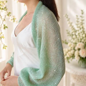 May include: A light mint green knitted bolero shrug featuring a sheer, open-weave texture with subtle integrated sequins that catch the light. The garment has long, voluminous sleeves with ribbed cuffs and a soft, airy drape over the shoulders.