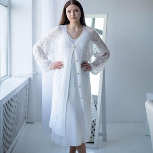 May include: A white, long-sleeved, open-front cardigan with a delicate lace pattern. The cardigan is worn over a white dress. The outfit is accessorized with brown sandals. The style is elegant and suitable for special occasions.