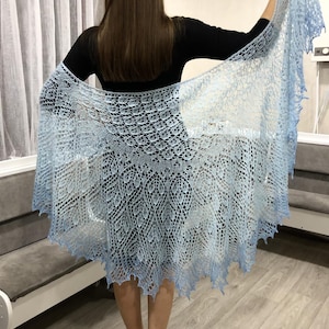 May include: A light blue knitted shawl with a delicate lace pattern. The shawl is draped over a person's shoulders and is held in place by their hands.