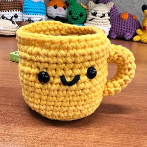 May include: A yellow crocheted mug with a smiling face and black eyes. The mug has a crocheted handle.
