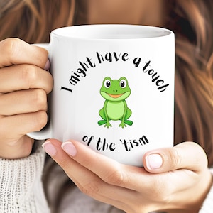 May include: White ceramic mug with the text "I might have a touch of the 'tism" and a cartoon frog illustration. The frog is green with a yellow belly and is centered on the mug. The mug has a standard handle.
