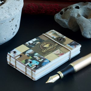 May include: A stack of Second Cup cafe cards bound with a light-coloured elastic band and a decorative metal gear. The cards feature images and the Second Cup logo. A gold fountain pen rests nearby.