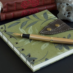 Op de afbeelding: Een gouden vulpen ligt op een groen notitieboek met een bladerenpatroon. Een bruine leren pennenhouder met messing knopen ligt naast de pen. De pagina's van het notitieboek zijn zichtbaar en de kaft is gestikt. Andere ledergebonden boeken zijn op de achtergrond.