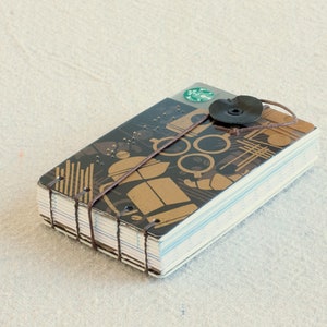 May include: A handmade journal with a Starbucks gift card cover. The cover has a brown and gold geometric design, a black button closure, and a brown string. The pages are white with blue edges.
