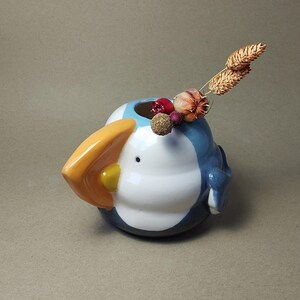 May include: A ceramic puffin-shaped vase. The vase has a large orange beak and a yellow spot. It is painted in white, blue, and black. The vase is decorated with dried flowers and berries. The vase has a small opening at the top.