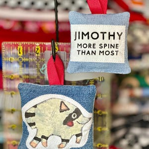 May include: Two square denim ornaments with red ribbon loops. One features a white patch with black text reading JIMOTHY MORE SPINE THAN MOST. The other displays a white patch with a stylized illustration of a raccoon with black and white stripes.