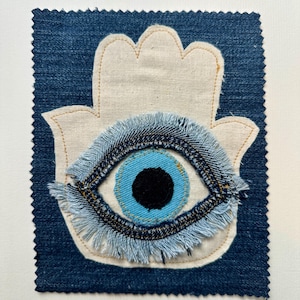 May include: A rectangular dark blue denim patch featuring a cream-colored fabric hamsa hand appliqué. Centered on the hamsa is a circular eye motif with a black pupil, a bright blue iris, and a frayed light blue denim fringe border.