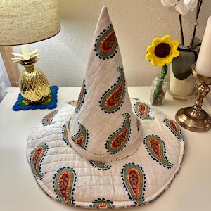 May include: A white quilted witch hat with a wide brim, adorned with a repeating paisley pattern in red, orange, blue, and green. The hat is set on a white surface, suggesting a decorative or costume item.
