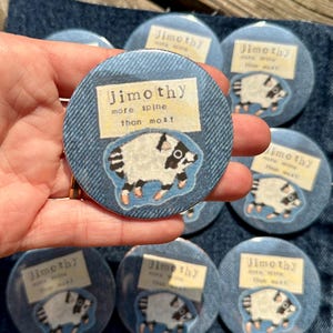 May include: A circular pinback button featuring a denim-textured background with a central illustration of a white and black opossum. Above the opossum is a rectangular label with the text "jimothy more spine than most" in black font.